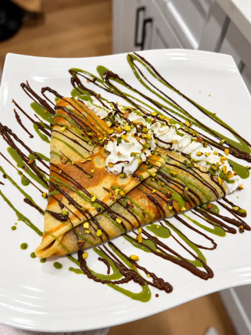 Folded Dubai chocolate crepe drizzled with dark chocolate and pistachio sauce, topped with whipped cream and pistachio crumbs